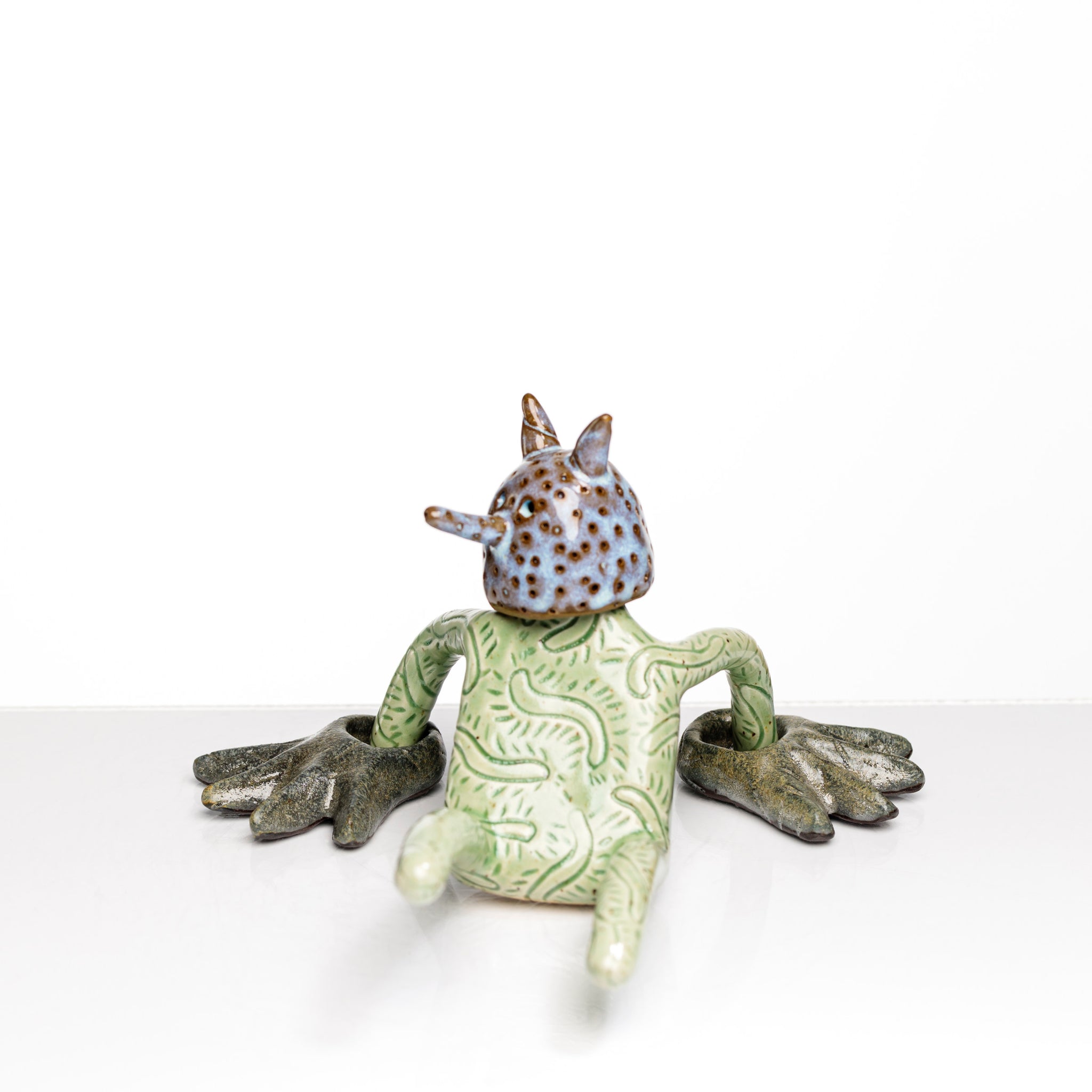 Sculpture en céramique par Fanny Mesnard. Un personnage vert avec une tête cornue et picotée est en perte d’équilibre sur le dos.