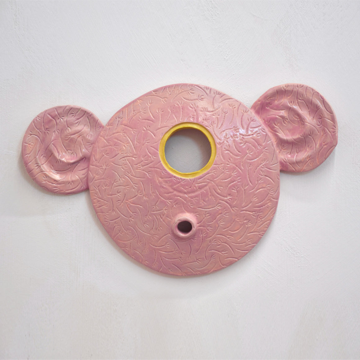 Masque mural en céramique de Fanny Mesnard. Un visage rose aux grandes oreilles avec un seul oeil jaune.