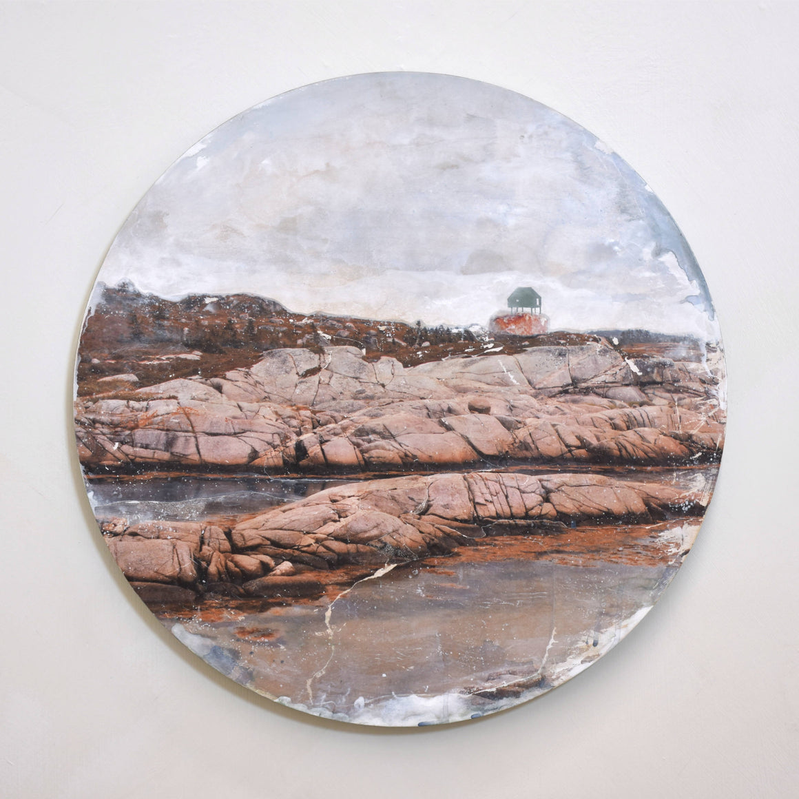 Tableau circulaire de l’artiste multidisciplinaire Caroline Bolieu. Une maison est posée, debout, sur des rochers en bord de mer.