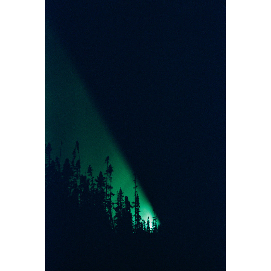 Un phare, la nuit, par Éva-Maude TC. Photographie en clair-obsur sur pellicule 35 mm. Lumière verte dans une nuit noire avec profil d’épinettes.