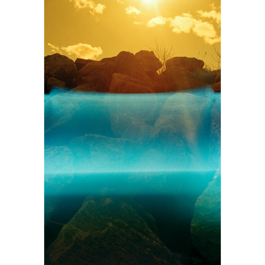 Zone intertidale, par Éva-Maude TC. Photographie contemporaine sur pellicule 35 mm, 24” x 36”, tirage limité de 4. Encadrement noir. Ciel jaune sur fleuve de lumière bleue, rochers de rivage.