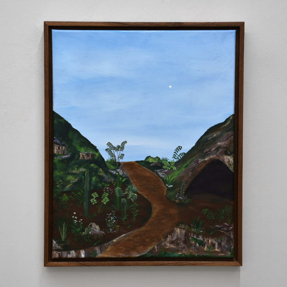 Sans titre, de Gabriel Rioux. Acrylique sur toile, 16” x 20”, cadre en noyer, 2022. Peinture de paysage, genre naïf, ciel bleu pâle et grotte sur colline verte.